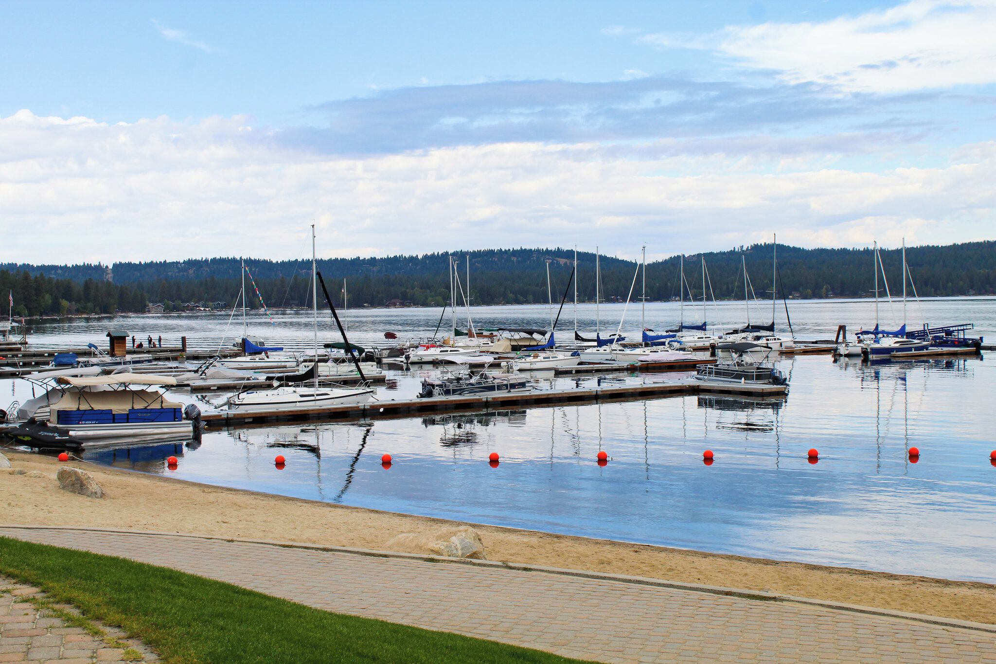 Eine Marina mit vertäuten Booten, ein sandiger Uferstreifen und ein bewaldeter Hügel im Hintergrund.