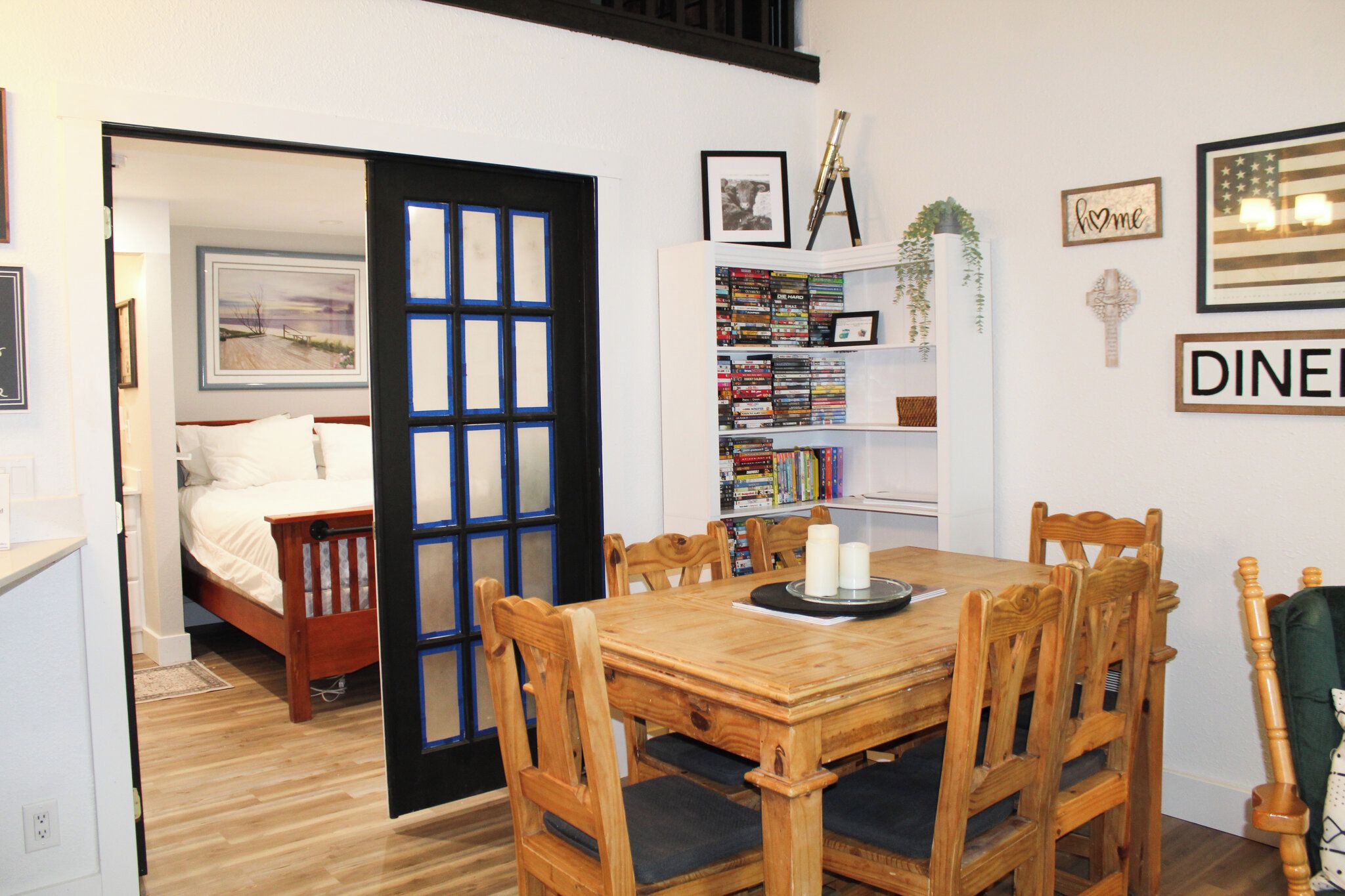 A room with a wooden dining table, chairs, a bookshelf, a bed, and a door leading to another room.
