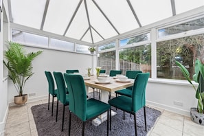 Une véranda avec une table à manger dressée pour six personnes, entourée de chaises vertes, et une grande plante en pot placée sur la gauche.