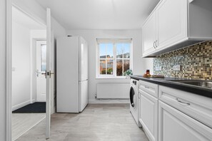 A modern kitchen with white cabinetry, a built-in washing machine, and a view through a window. - House Sleeps 9 with Enclosed Garden (Solihull)