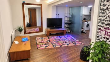 A living room with a wooden floor, a TV on a stand, a patterned rug, a desk with a laptop, and a mirror.