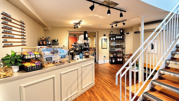 A café with a counter displaying various coffee options and pastries, a staircase, and a shelving unit with beverages.