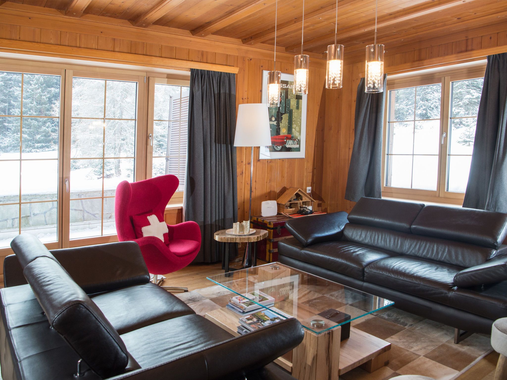 A modern living room with a glass coffee table, leather sofas, a red chair, and large windows with curtains.