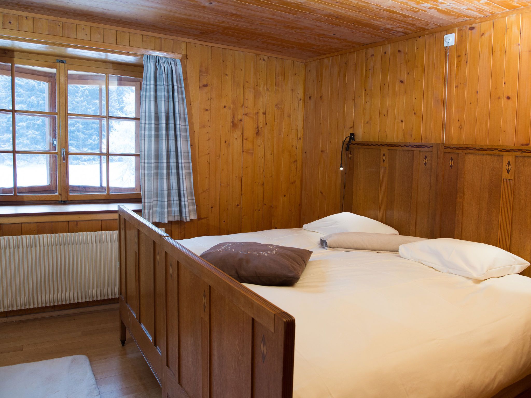 A wooden cabin room with a bed, a window with curtains, and a radiator.