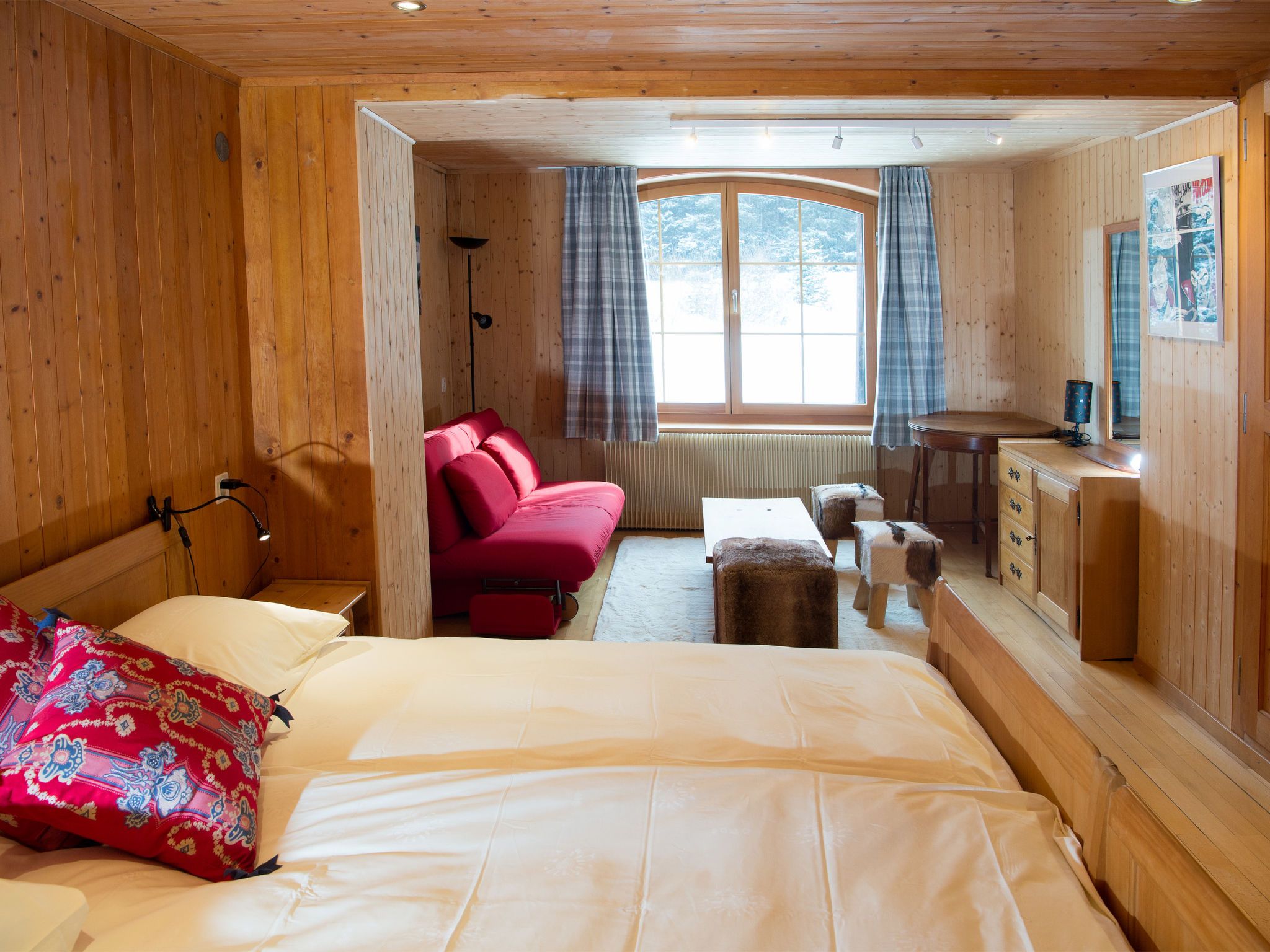 Una habitación de cabaña de madera con una cama, un sofá rojo, una mesita y una ventana con cortinas.