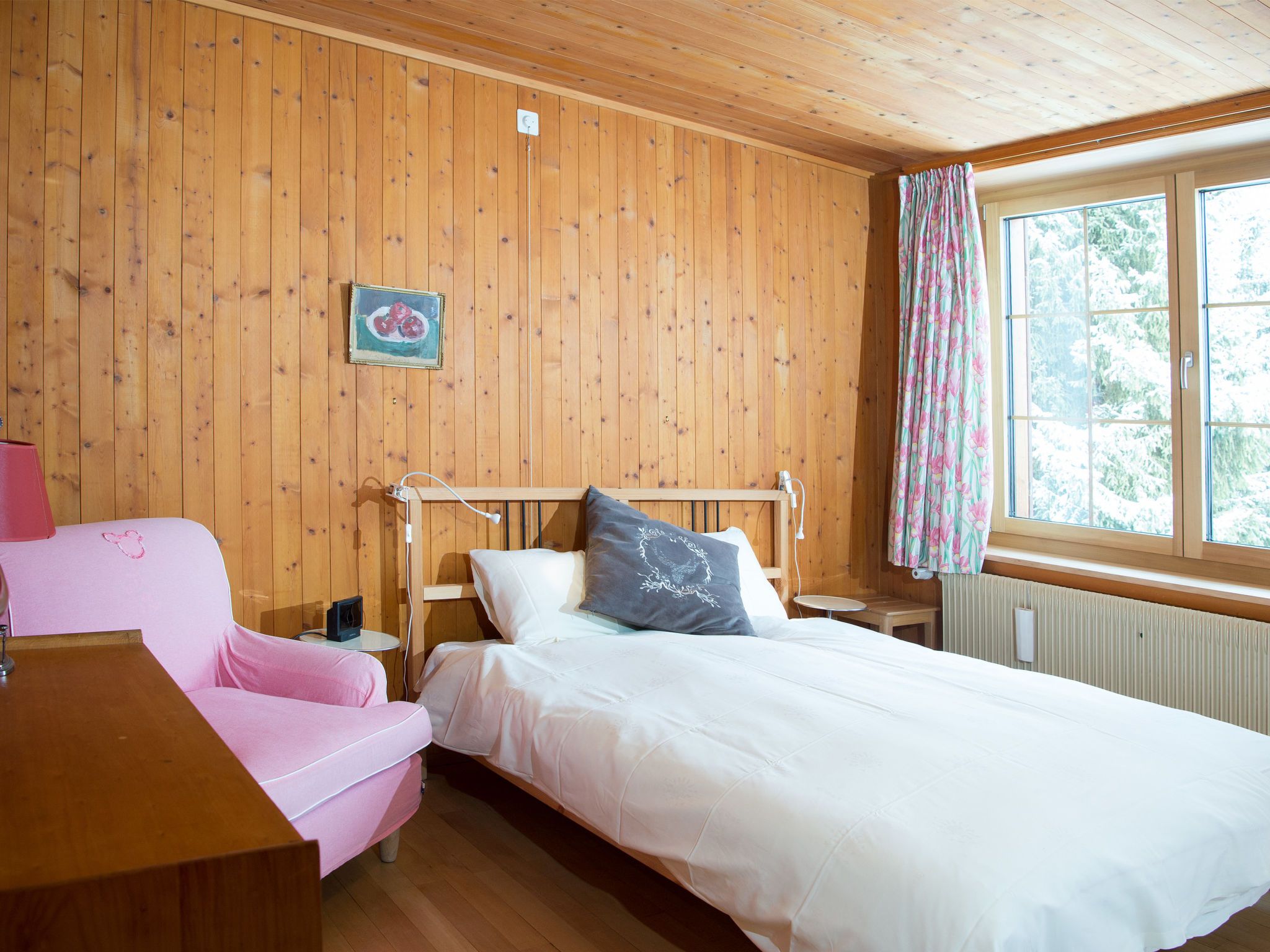 Ett rum med träpanel på väggarna, en säng, en rosa stol och ett litet bord med en lampa.