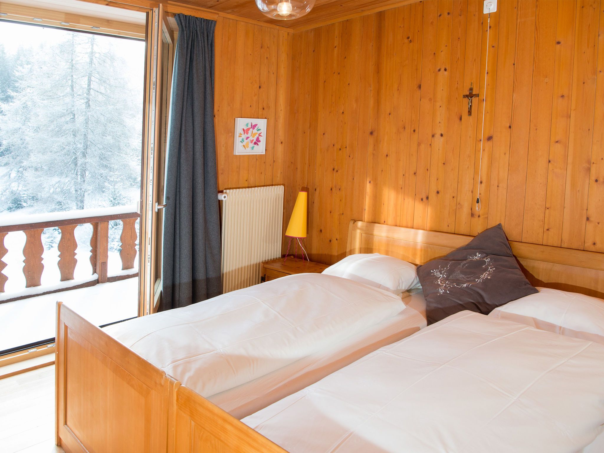 Habitación de cabaña de madera con una cama, una ventana con cortinas, un radiador y una lámpara fijada a la pared.