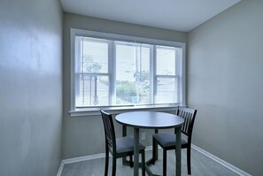 Ein Zimmer mit einem runden Tisch und vier Stühlen, einem Fenster mit Jalousien und Blick auf ein Gebäude.