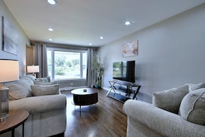 A modern living room with a sofa set, a coffee table, a flat-screen TV, and a large window providing a view of the outdoors. - Your Chicago Home Between Homes - 4BR Furnished Home (Chicago)