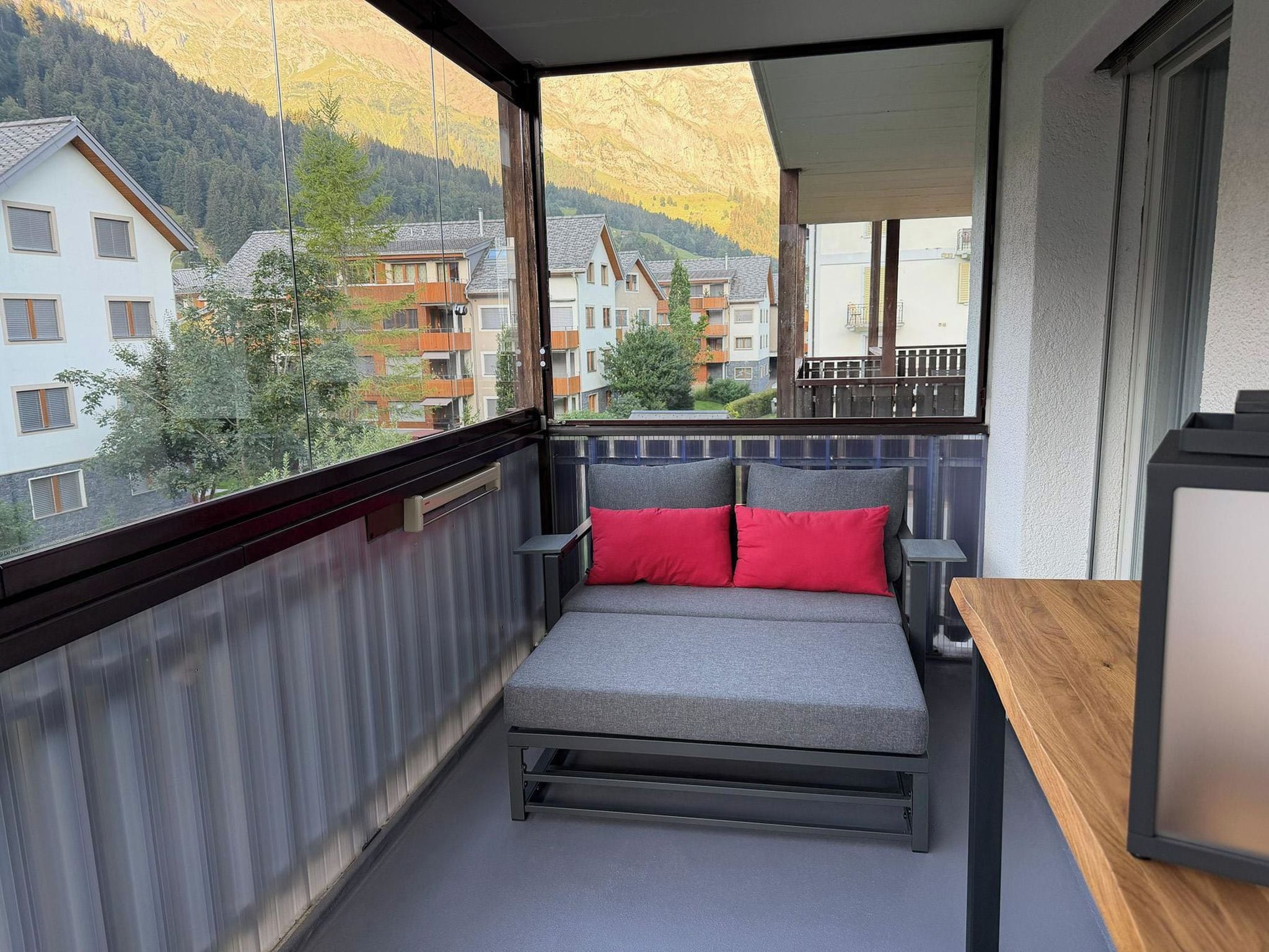 A balcony with a grey sofa and red cushions, overlooking a residential area with houses and trees.