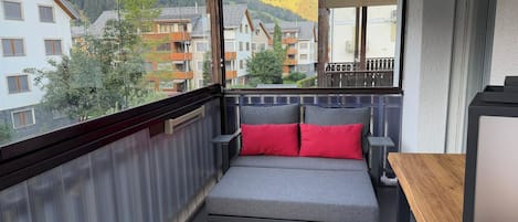 A balcony with a grey sofa and red cushions, overlooking a residential area with houses and trees.