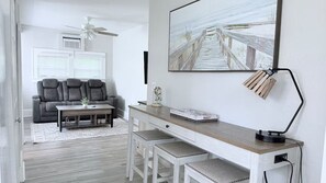 Una sala moderna con un sofá gris, una mesa de centro de madera y un escritorio con una lámpara. Un cuadro grande de un muelle de madera adorna la pared.