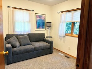 A room with a grey sofa, a framed abstract painting, a floor lamp, and a window with a view of trees. - Cozy 2-bedroom house in a woodsy setting, near restaurants, shops, and more. (Pontiac)