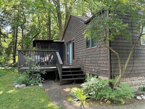 A wooden cabin with a deck and a small porch, surrounded by a garden and trees. - Cozy 2-bedroom house in a woodsy setting, near restaurants, shops, and more. (Pontiac)