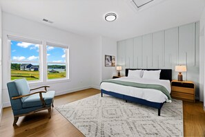 A bedroom with a large bed, two bedside lamps, a chair, and a view of a house through two windows. - Lakefront Lake Anna | Theater, Hot Tub, Beach (Mineral)
