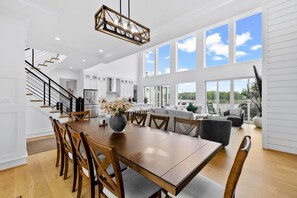 A spacious dining area with a wooden table and chairs, a kitchen in the background, and large windows offering a view of the outdoors. - Lakefront Lake Anna | Theater, Hot Tub, Beach (Mineral)