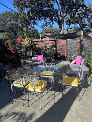 Un patio al aire libre con una mesa de vidrio y sillas de metal, rodeado por una cerca de madera y plantas en flor.