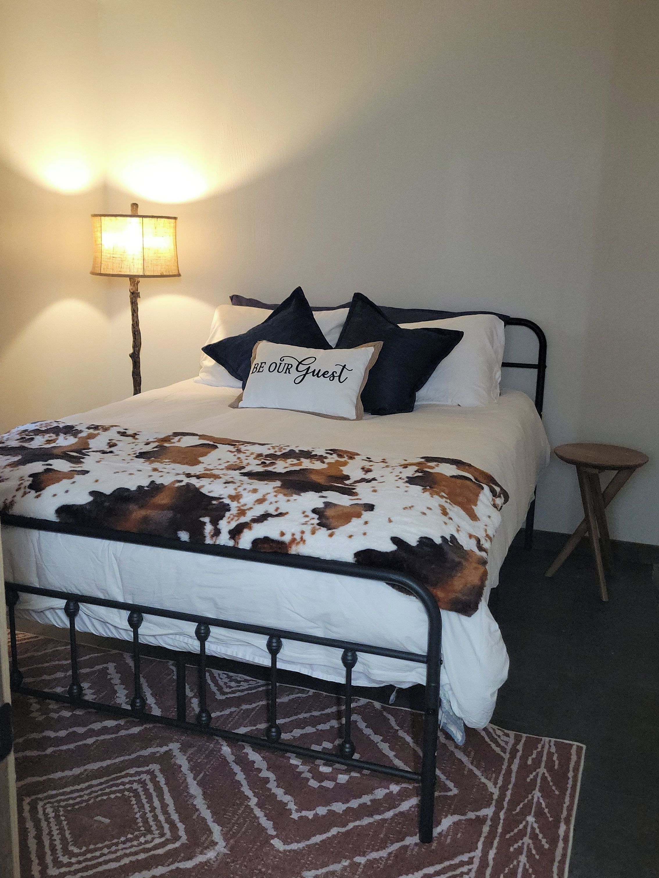 Een bed met een deken met koeienprint, een kussen met de tekst 'BE OUR GUEST', een lamp en een klein krukje.