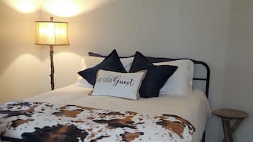 Een bed met een deken met koeienprint, een kussen met de tekst 'BE OUR GUEST', een lamp en een klein krukje.