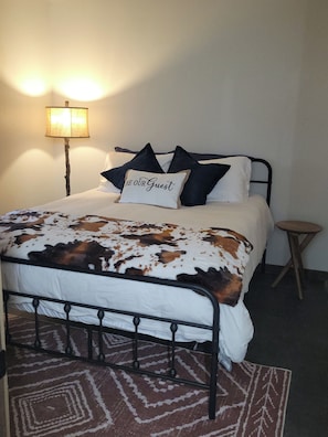 A bed with a cow print blanket, a pillow with 'BE OUR GUEST' text, a lamp, and a small stool.