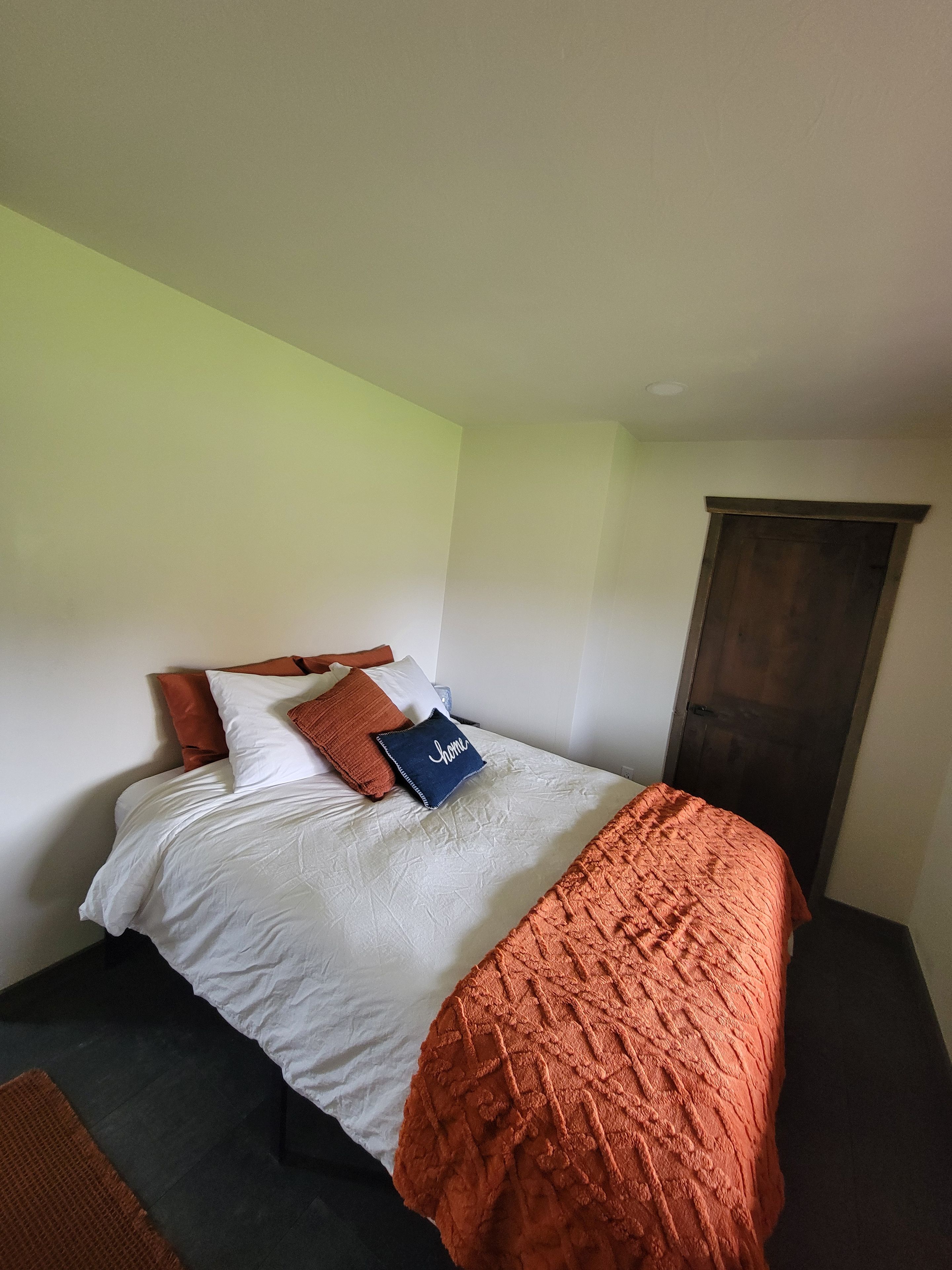 A single bed with a white mattress, a brown blanket, and several pillows in a room with a plain wall and a wooden door.
