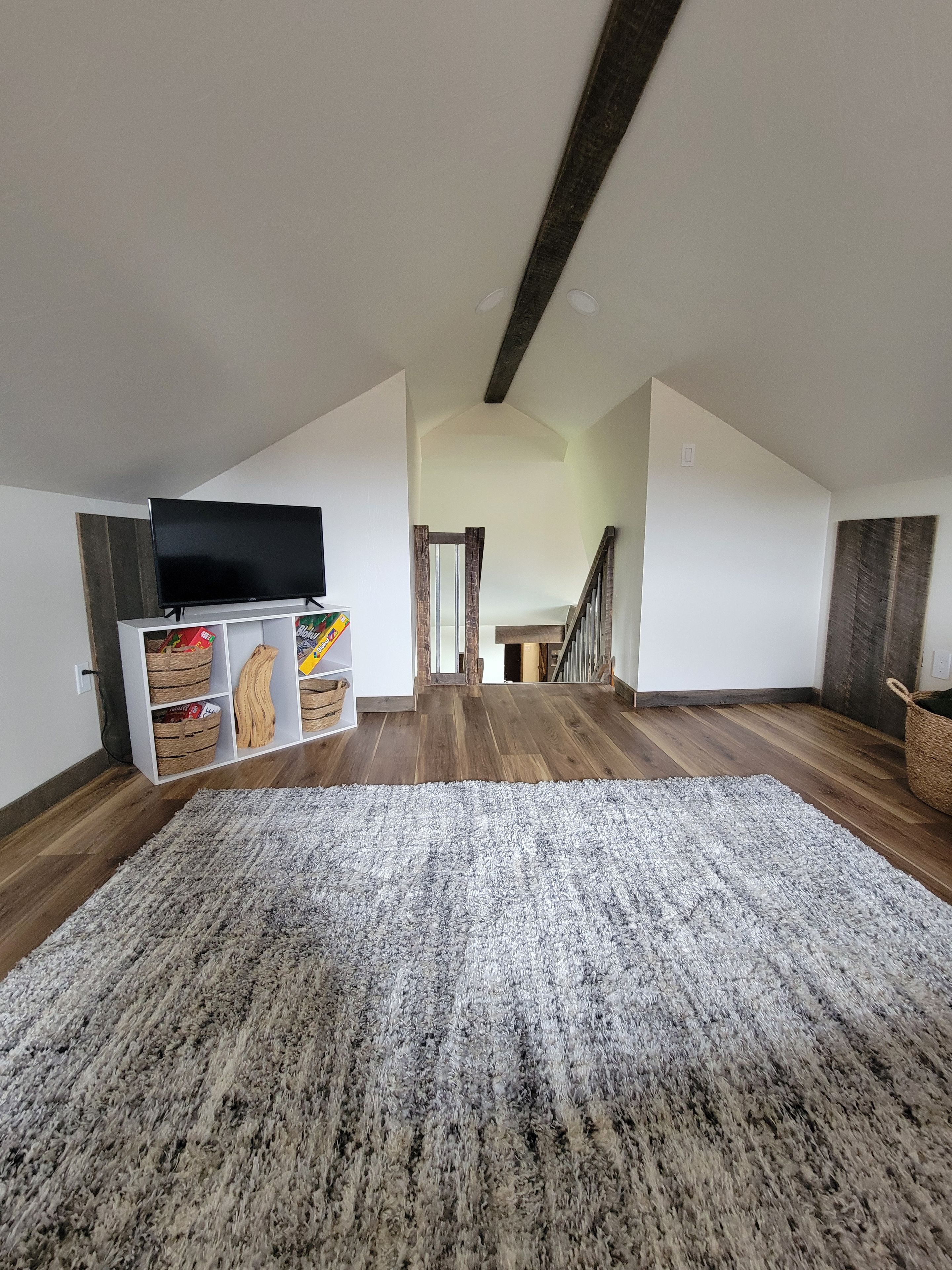 A spacious room with a large grey rug, wooden flooring, a television on a white stand, and a staircase leading to an upper level.
