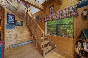 A wooden interior with a staircase, a window with a view of greenery, and a bookshelf with books.