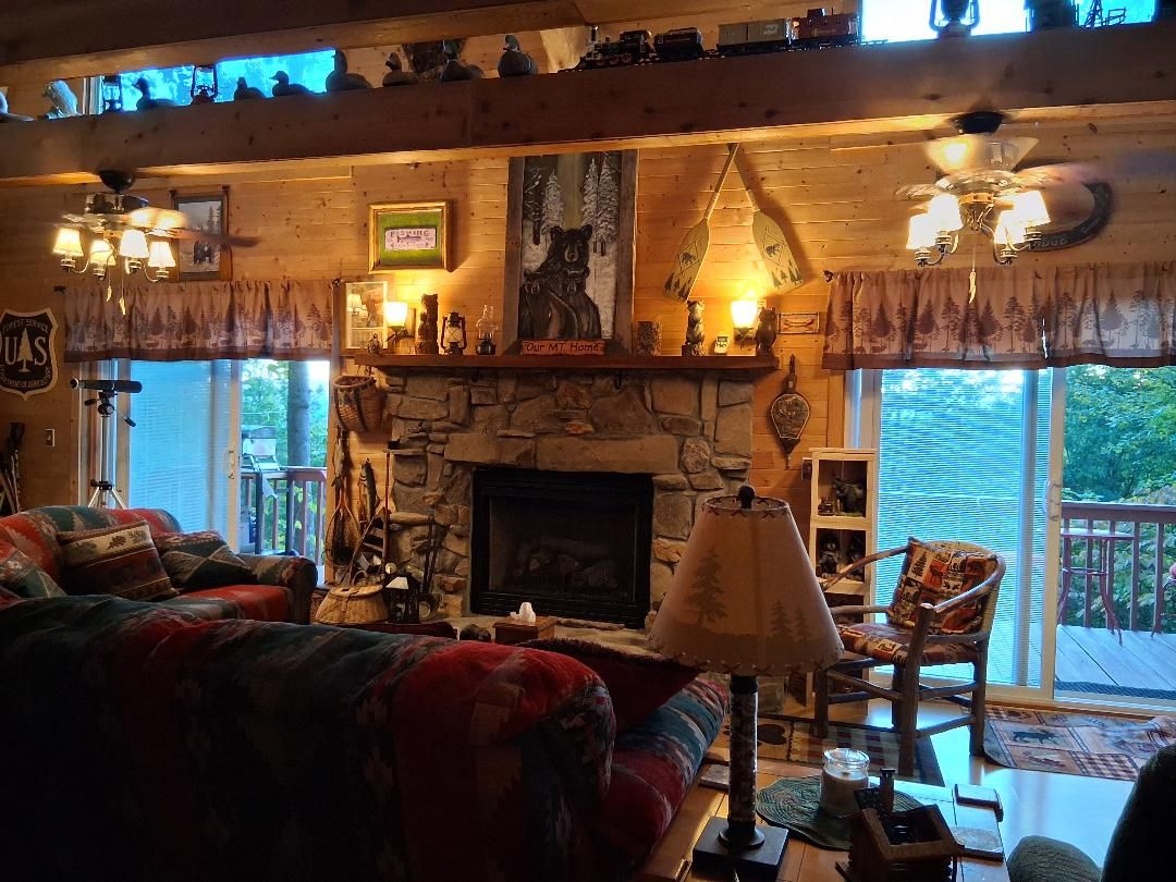 A cozy living room with a stone fireplace, a ceiling fan, and a large window overlooking a deck.