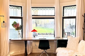 A room with a desk, chair, and a sofa. There are three windows with curtains, a lamp, and a vase with flowers on the desk. - New Modern Craftsman Guest House with Bay Windows (San Jose)