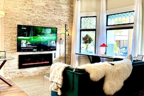 A living room with a flat-screen TV mounted on a brick wall, a fireplace below it, a green sofa with a white fur throw, and a window with a view of greenery. - New Modern Craftsman Guest House with Bay Windows (San Jose)