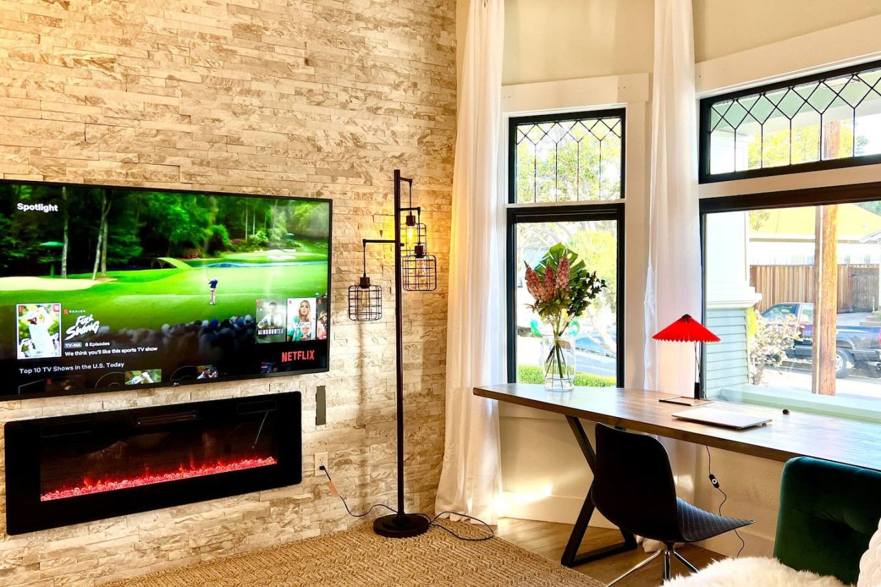 A modern living room with a flat-screen TV mounted on a brick wall, a wooden desk with a chair, and a large window with a view of the outdoors.
