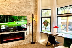 A modern living room with a flat-screen TV mounted on a brick wall, a wooden desk with a chair, and a large window with a view of the outdoors. - New Modern Craftsman Guest House with Bay Windows (San Jose)
