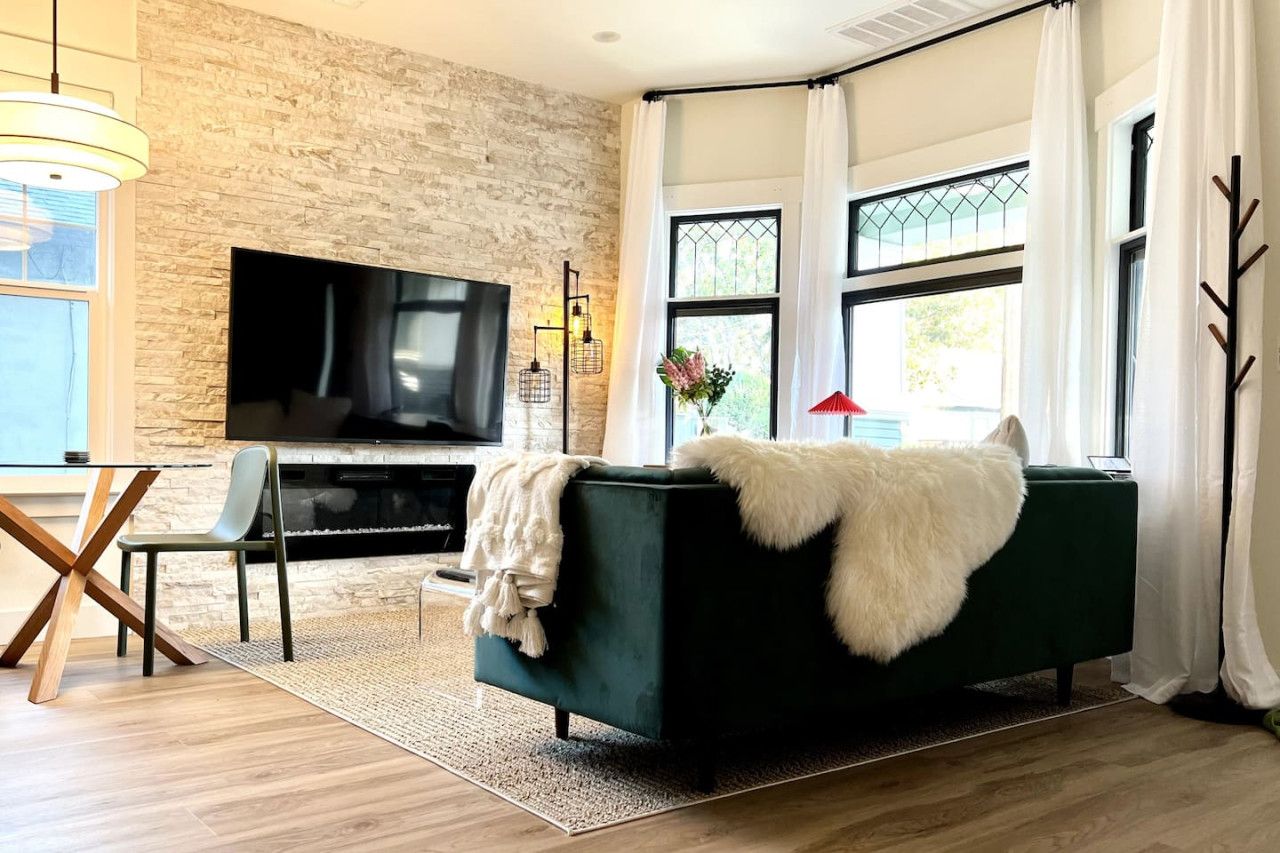 A modern living room with a stone wall, a flat-screen TV, a dark green sofa, and a dining area with a glass table and chairs.