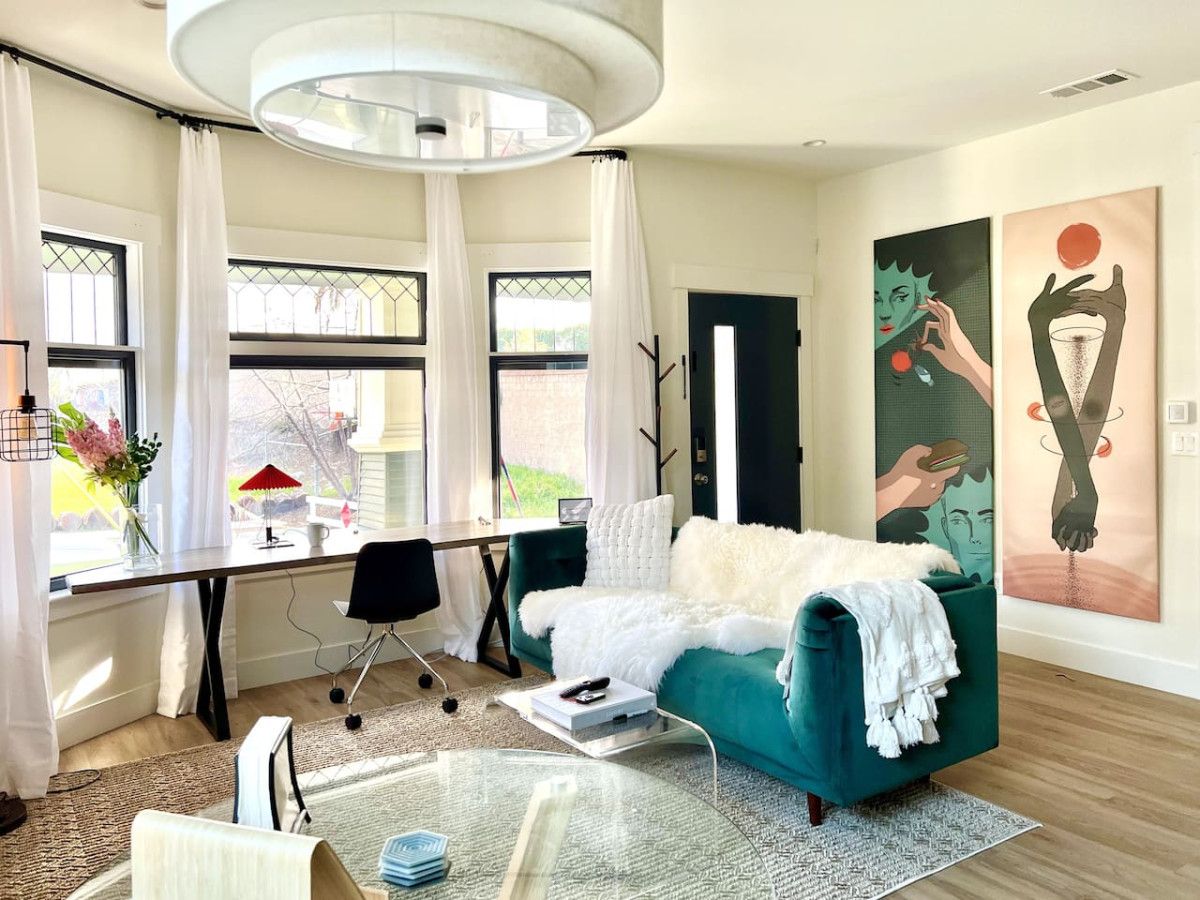 A modern living room with a teal sofa, a glass coffee table, a desk with a chair, and artwork on the wall.