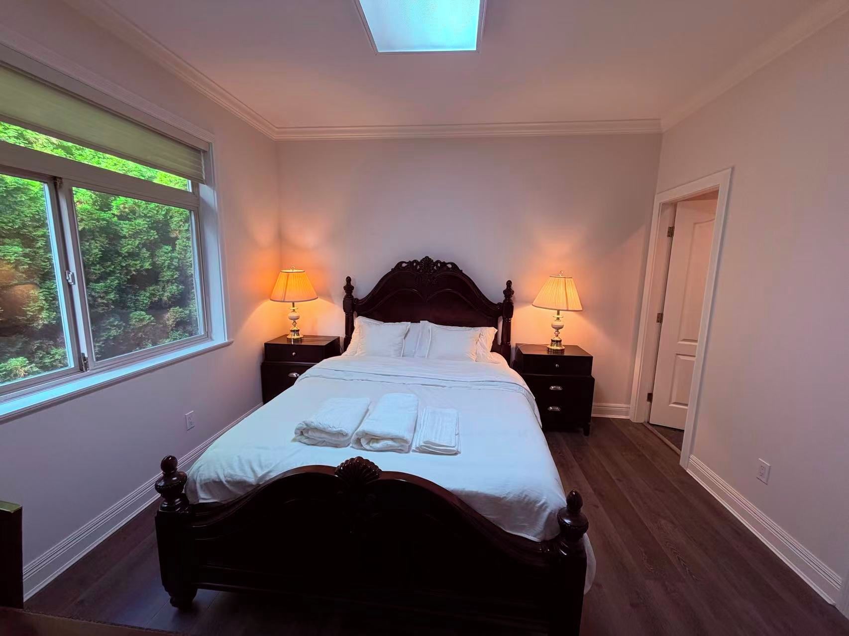 A neatly made bed with white linens and two bedside tables with lamps, a skylight above, and a view of greenery through the window.