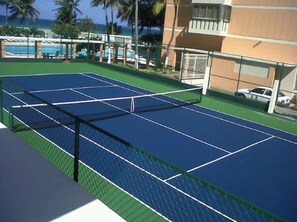 An outdoor tennis court with a net, surrounded by a fence, and a building with a gated entrance in the background. - Gorgeous Condado 3-Bedroom + 2 Bath w/ OceanView! (San Juan)