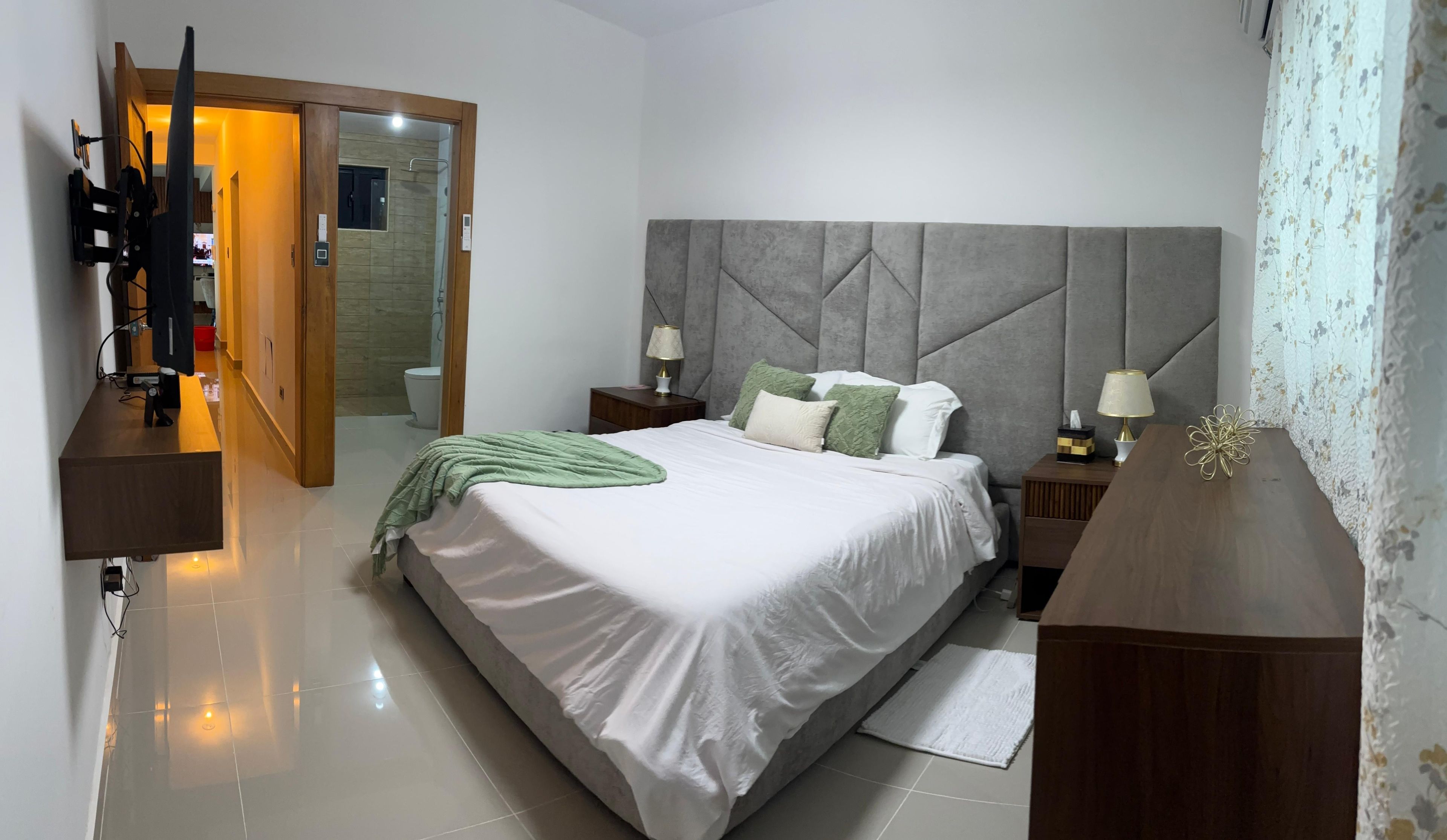 Un dormitorio moderno con una cama grande, mesitas de noche, un armario y un baño visible a través de una puerta abierta.
