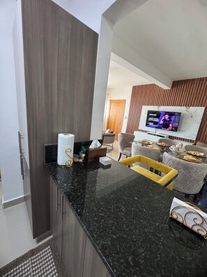 A modern kitchen with a marble countertop, wooden cabinets, and a dining area with a TV displaying a show. - Spacious 3-bedroom apartment in peaceful Santiago de los Caballeros with AC (Santiago de los Caballeros)