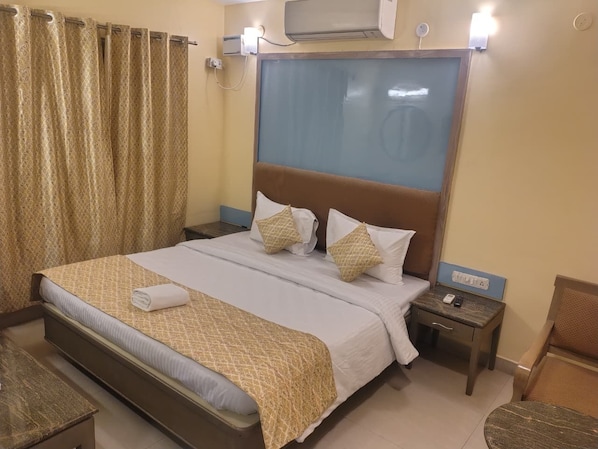 A hotel room with a bed, bedside tables, a nightstand, an air conditioning unit, and a window with curtains.