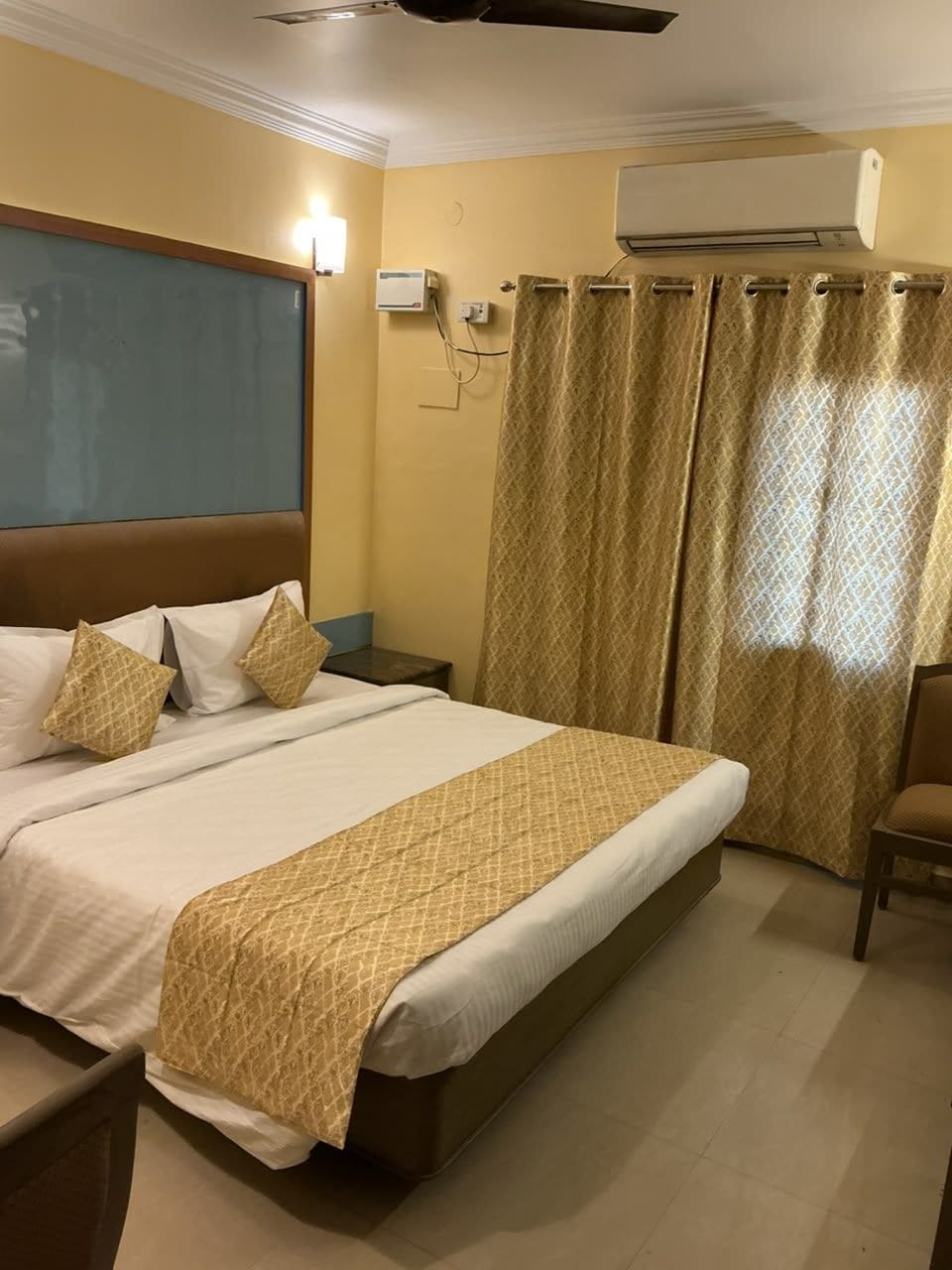 Quarto de hotel com cama, mesa de cabeceira, cadeira, aparelho de ar condicionado e cortina.