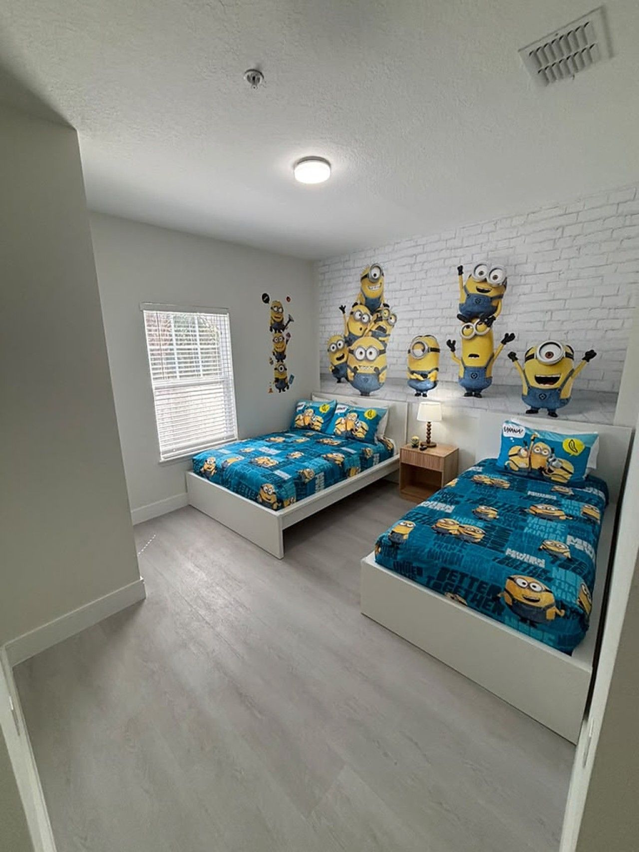 A bedroom with two beds, a window with blinds, a white brick wall, and a ceiling light.