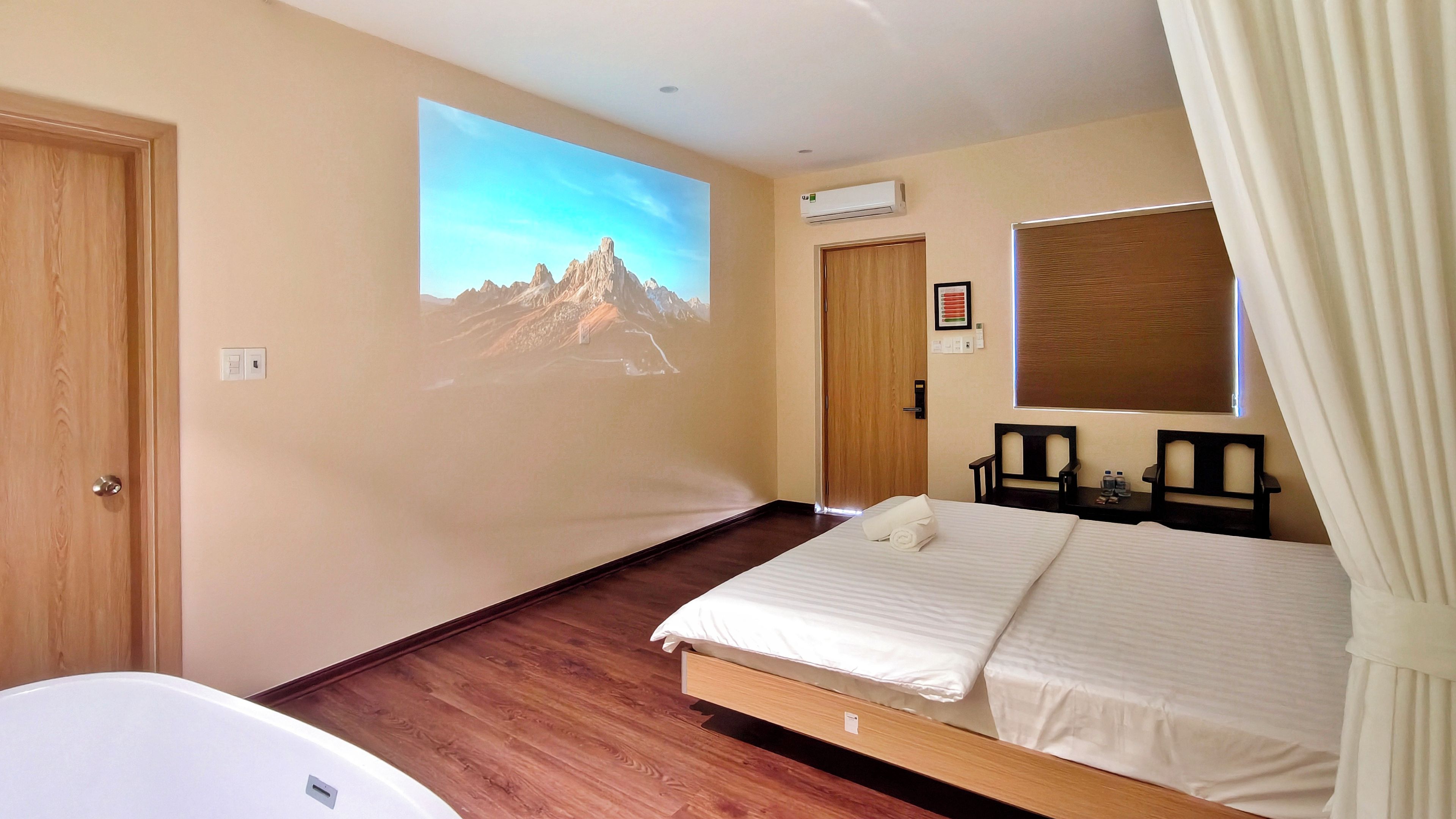 A hotel room with a bed, a wall-mounted projector, a window with curtains, a wooden door, and a small table with a chair.
