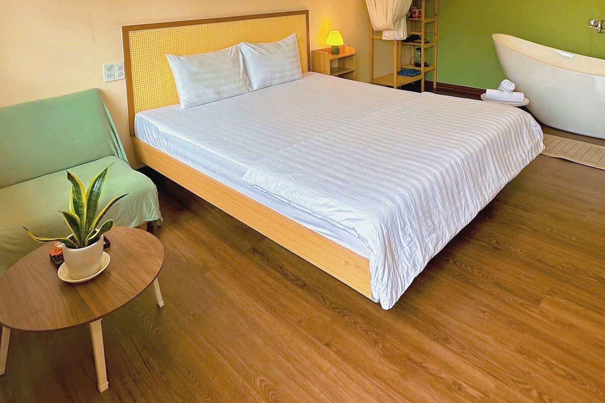 Habitación de hotel con una cama, un sillón verde, una mesita con una planta, una lámpara de noche y una bañera.