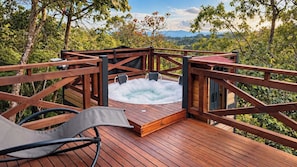 Um deck de madeira com hidromassagem, cercado por guarda-corpo de madeira, com vista para uma paisagem arborizada.