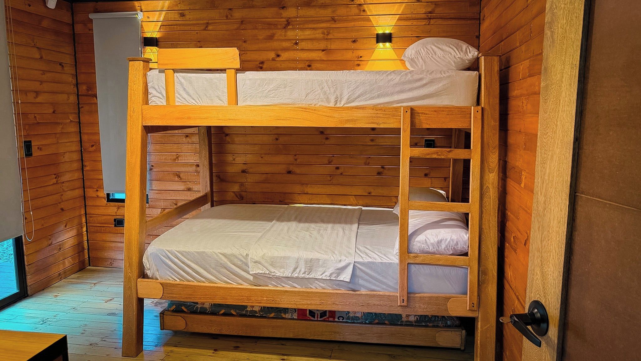 A bunk bed with a wooden frame and white bedding, set in a room with wooden walls and a door.