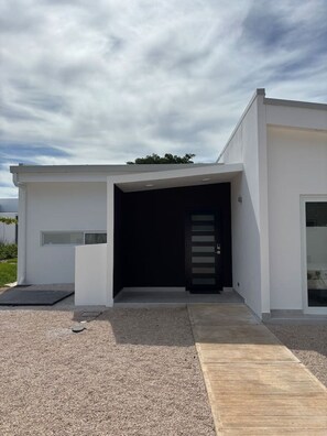 A modern building with a flat roof, a black entrance door, and a gravel driveway leading to a wooden ramp. - Serene, Contemporary Retreat with Plunge Pool (Playa Avellanas)