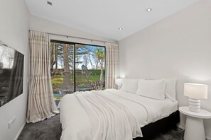 Ein Schlafzimmer mit einem großen Bett, einem Fernseher, einem Nachttisch mit Lampe und Blick auf einen Garten durch eine Schiebetür.