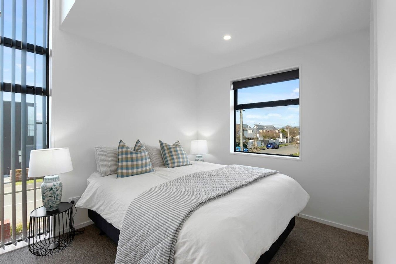 A bedroom with a large bed, checkered pillows, a bedside table with a lamp, and a window with a view of a street.