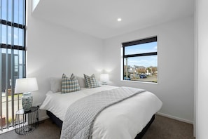 A bedroom with a large bed, checkered pillows, a bedside table with a lamp, and a window with a view of a street. - Modern Merivale Gem | Flexible Beds & Parking (Christchurch)