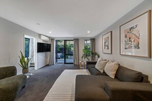 A modern living room with a sofa, a coffee table, and a TV. There is a sliding glass door leading to an outdoor area. - Modern CBD retreat with parking and courtyard (Christchurch)